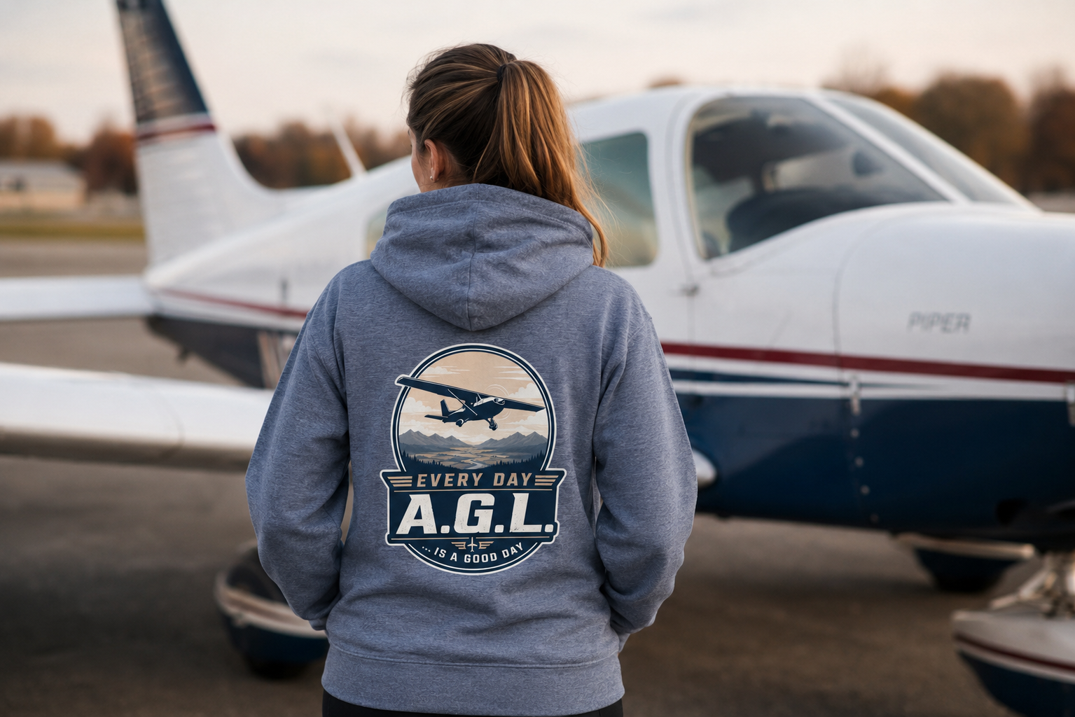A pilot on the ramp wearing the Every Day A.G.L. hoodie beside a Piper